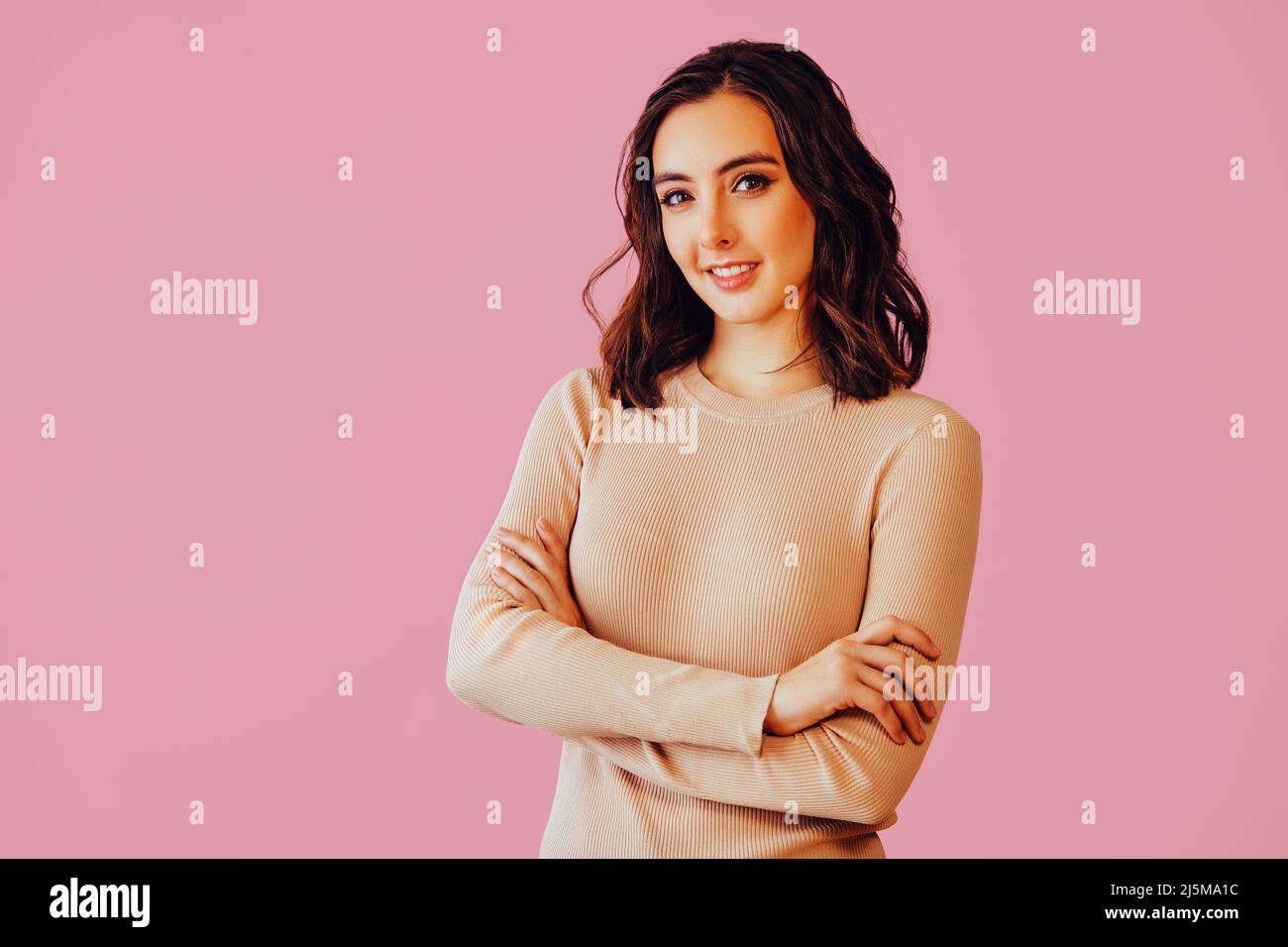 Belle confiante brune jeune femme souriant regardant la caméra avec les bras croisés. Espace de copie arrière-plan rose portrait de studio Banque D'Images