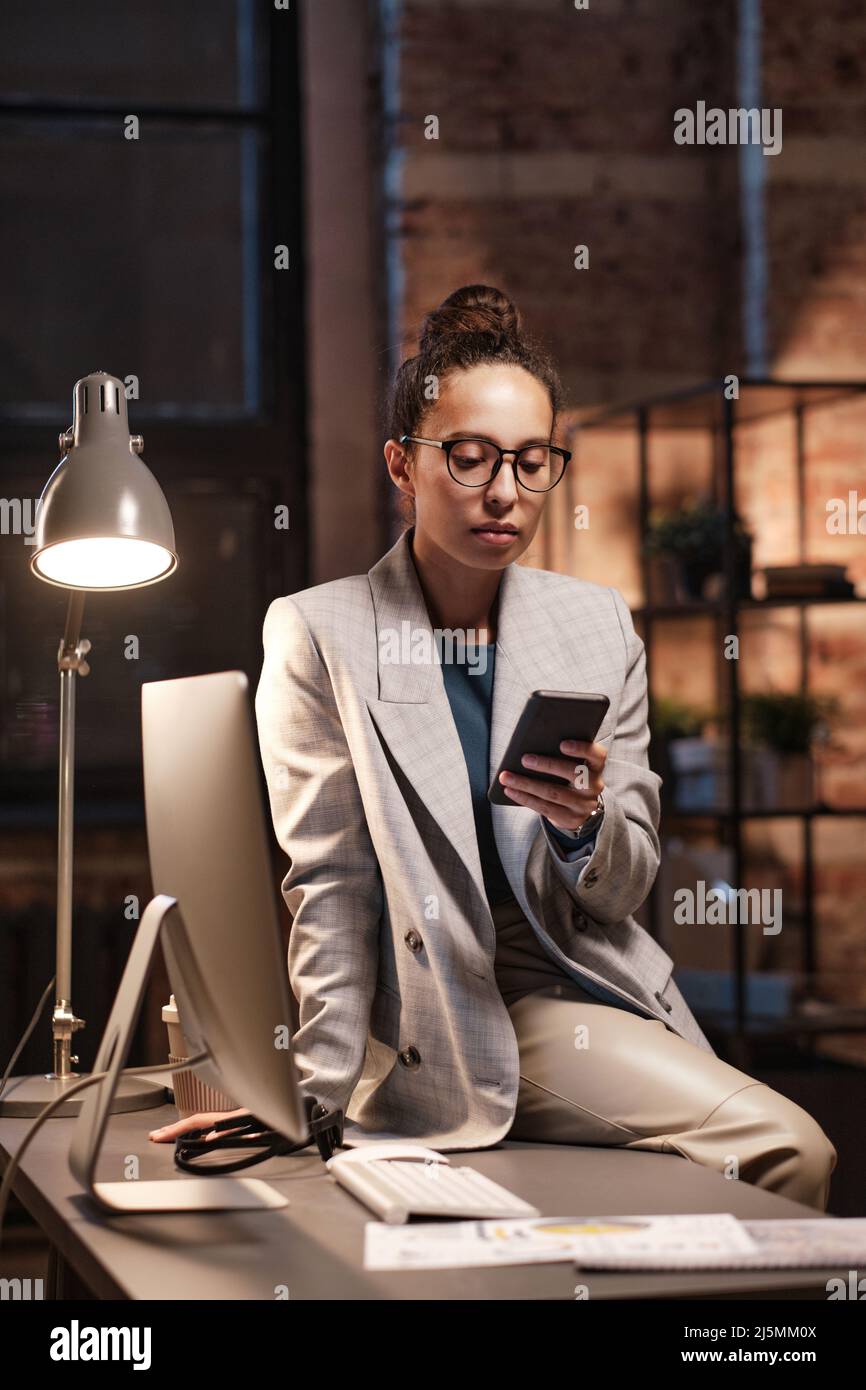 Jeune femme d'affaires sérieuse de race mixte dans des lunettes assis sur une table avec ordinateur et défilant dans les médias sociaux dans le bureau sombre Banque D'Images
