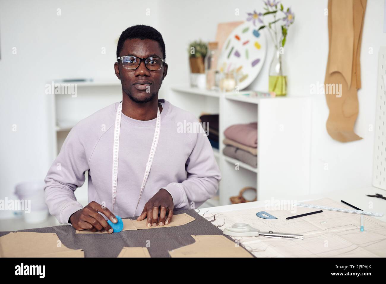 Portrait du jeune Noir tailleur assis à une grande table dans son studio et décrivant et coupant les détails du vêtement Banque D'Images