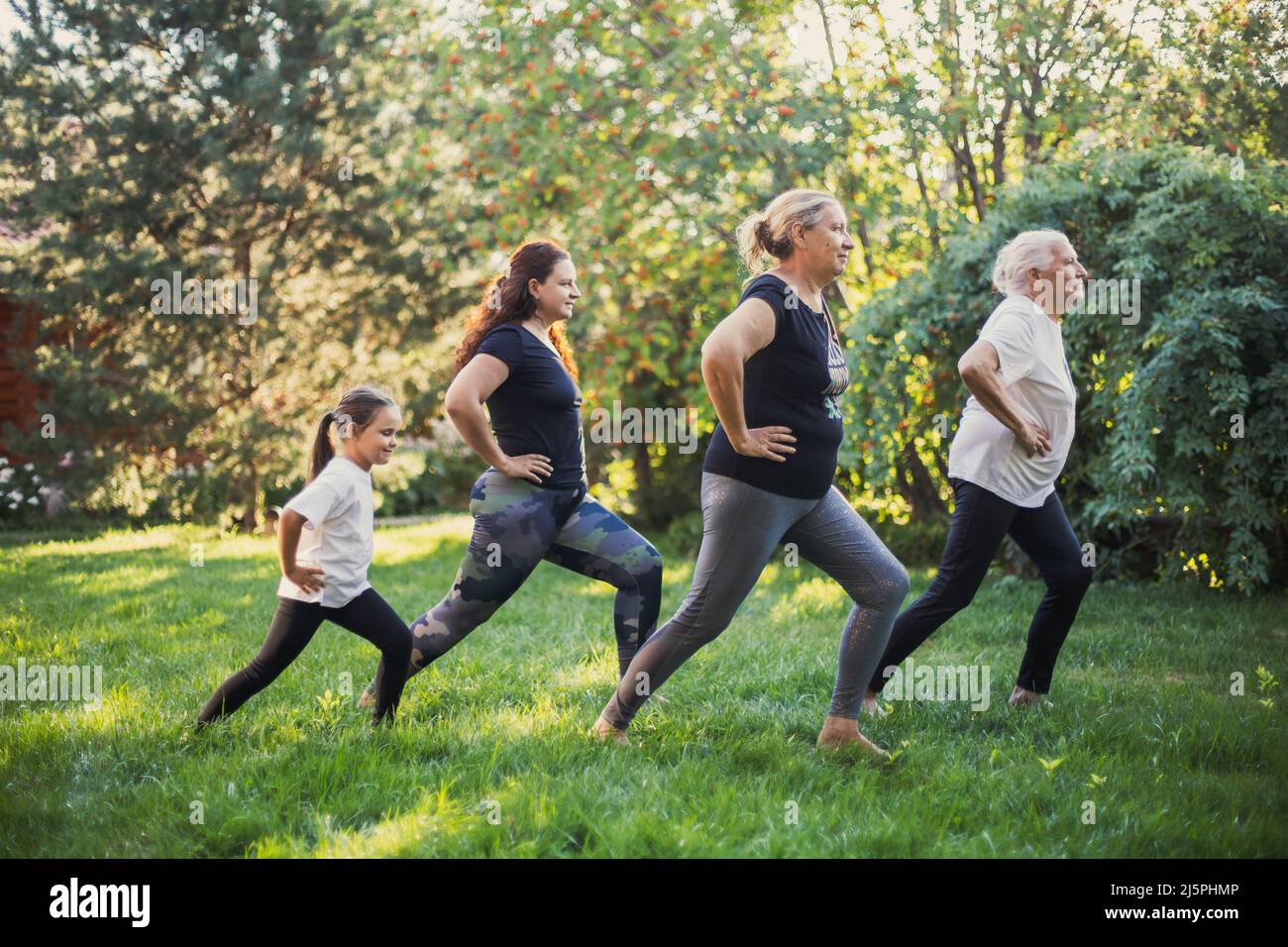Quatre femelles de différentes générations de famille faisant des exercices physiques pour étirer les jambes ensemble sur la cour pleine d'herbe verte et d'arbres Banque D'Images