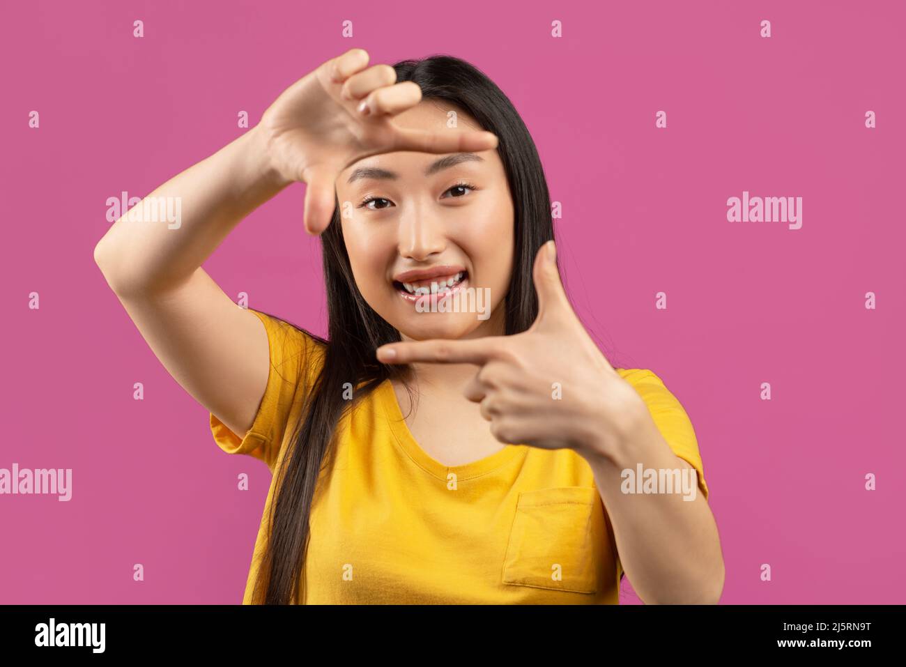Jolie femme asiatique regardant à travers les doigts comme l'appareil photo, posant sur fond violet, faisant photo cadre d'appareil photo Banque D'Images