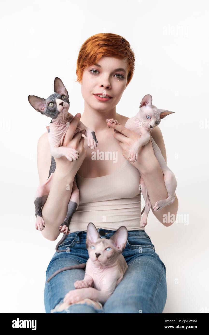 REDHEAD jeune femme tenant deux chats Sphynx dans les mains et un chaton allongé sur ses jambes. Jolie femme avec des cheveux courts en T-shirt et Jean regardant l'appareil photo Banque D'Images