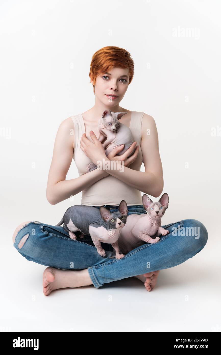 REDHEAD jeune femme tenant le chat de Sphynx entre les mains et deux chatons assis sur ses jambes. Femme taille basse avec cheveux courts en T-shirt et Jean regardant l'appareil photo Banque D'Images