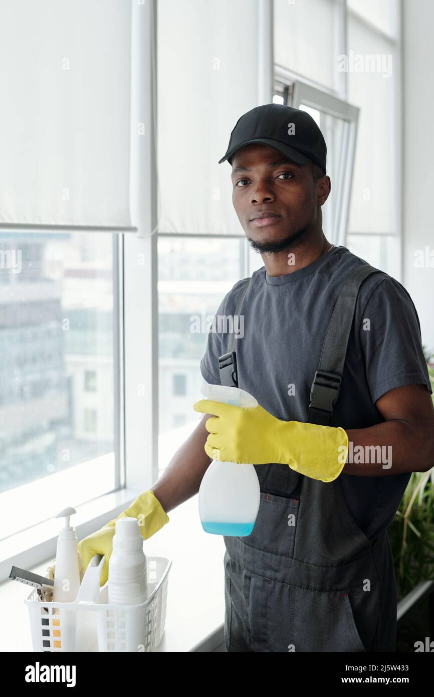 Jeune Africain américain contemporain avec détergent en bouteille plastique debout près du seuil avec récipient tout en lavant les fenêtres Banque D'Images