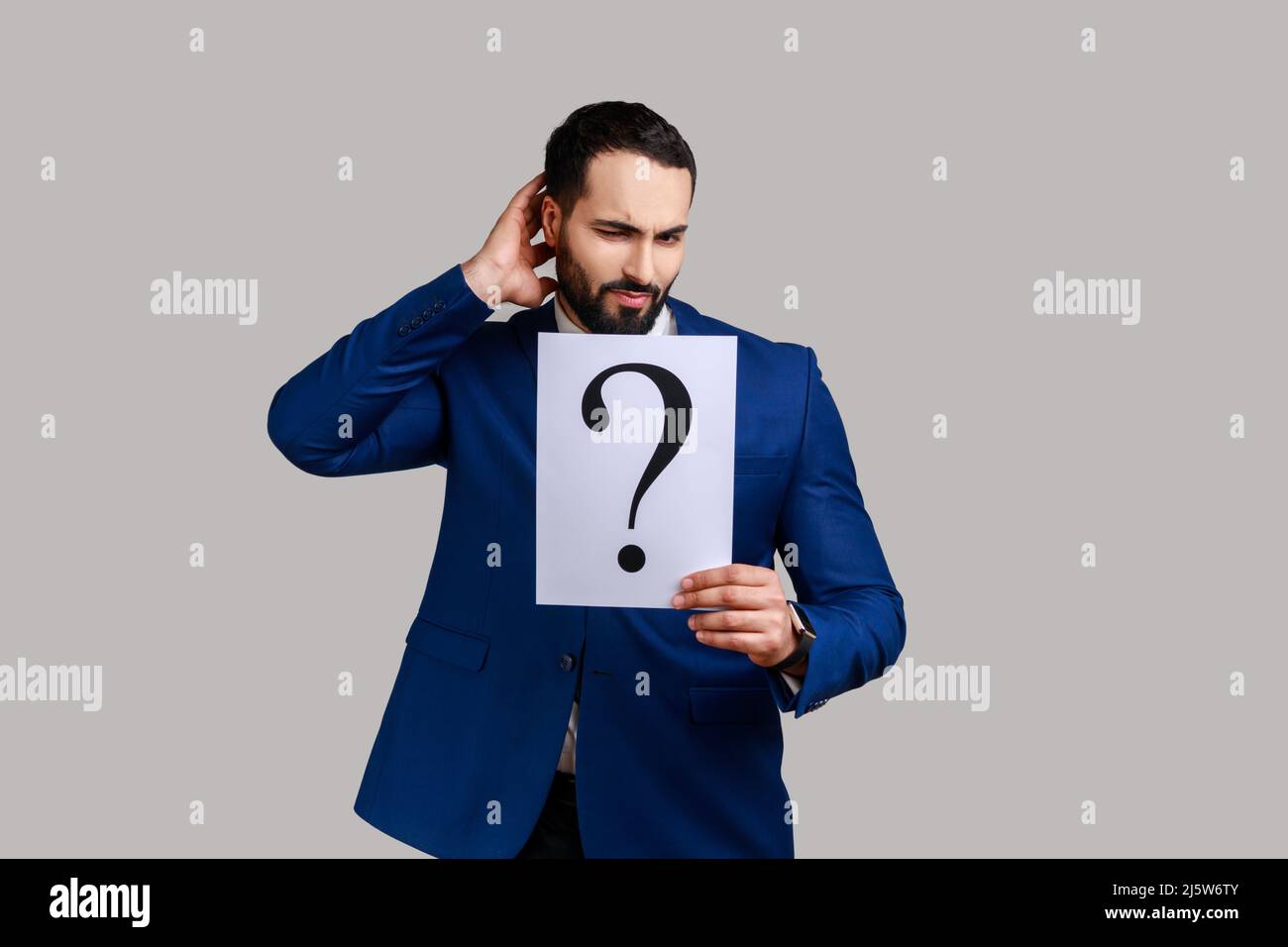Homme barbu et pensif tenant un point d'interrogation, trouvant une solution intelligente, demandant des conseils, regardant un appareil photo, portant un costume officiel. Prise de vue en studio isolée sur fond gris. Banque D'Images