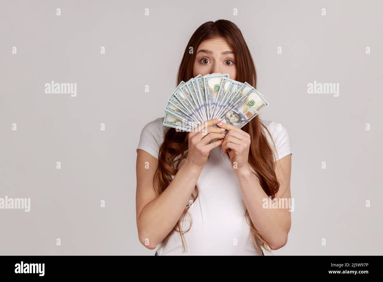 Surprise heureuse femme avec des cheveux bruns cache face derrière un fan de billets de dollars, retrait d'argent sans intérêt, portant un T-shirt blanc. Prise de vue en studio isolée sur fond gris. Banque D'Images