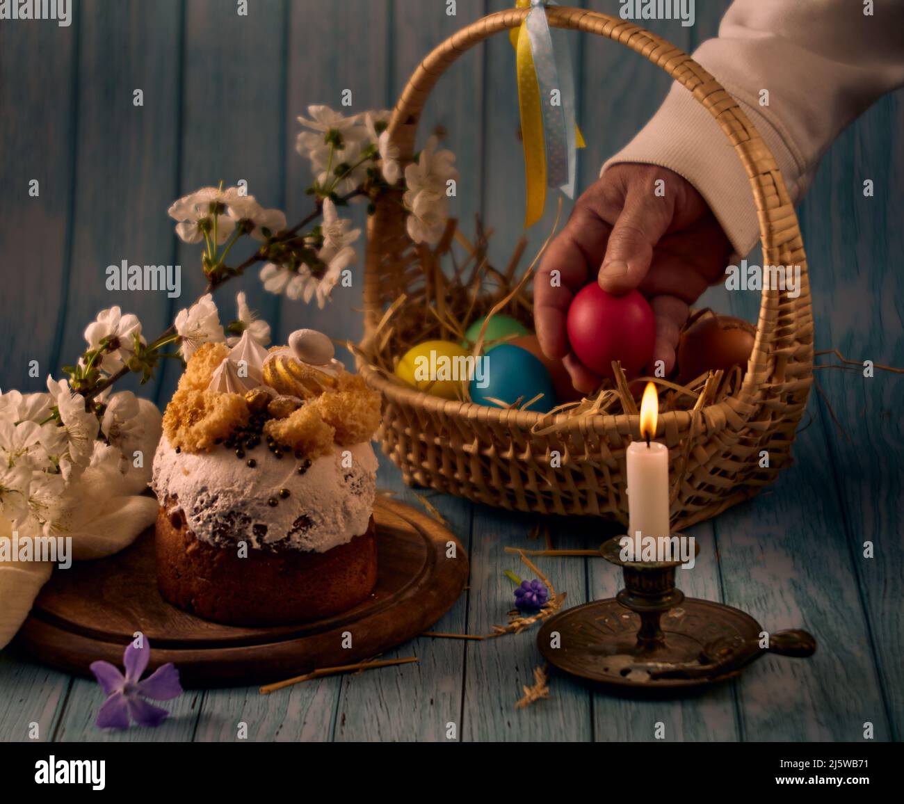 Gâteau de Pâques, un panier avec des oeufs colorés, un chandelier de bronze avec une bougie en feu, une main tenant un oeuf et des fleurs de printemps avec un backgroun flou Banque D'Images