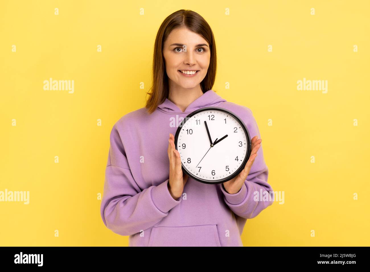 Portrait d'une femme heureuse positive tenant une grande horloge murale, regardant l'appareil photo avec un sourire agréable, le temps d'aller, portant un pull à capuche violet. Studio d'intérieur isolé sur fond jaune. Banque D'Images