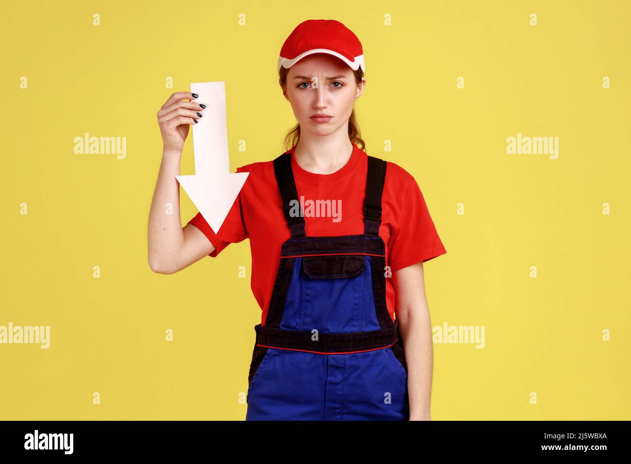 Portrait d'une femme travailleur triste contrariée debout avec une flèche blanche dans les mains indiquant vers le bas, regardant l'appareil photo, portant une combinaison et une casquette rouge. Studio d'intérieur isolé sur fond jaune. Banque D'Images