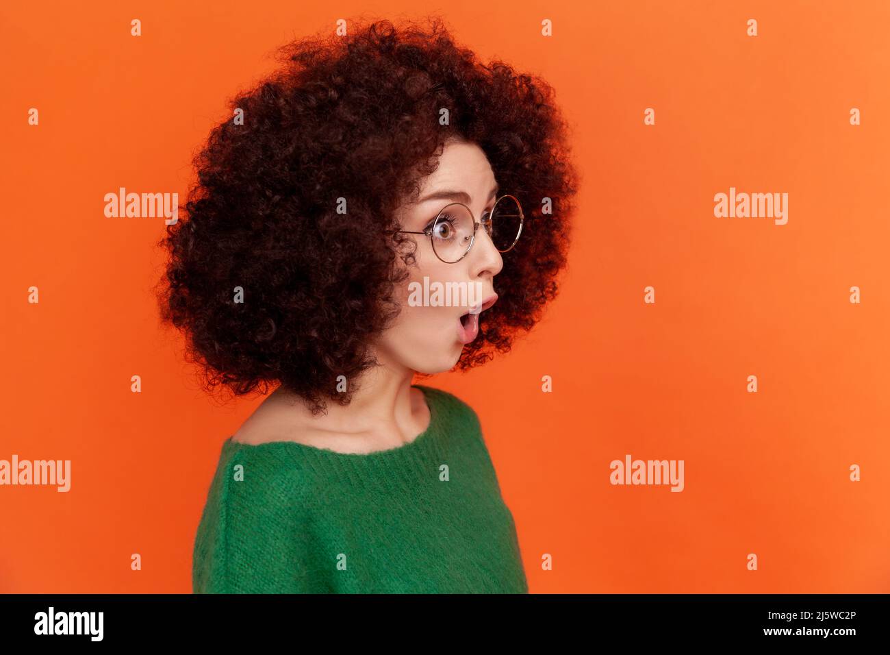 Portrait de profil de la femme choquée avec la coiffure afro en vert décontracté style chandail debout avec la bouche ouverte, regardant loin, voit quelque chose de surprenant. Studio d'intérieur isolé sur fond orange. Banque D'Images