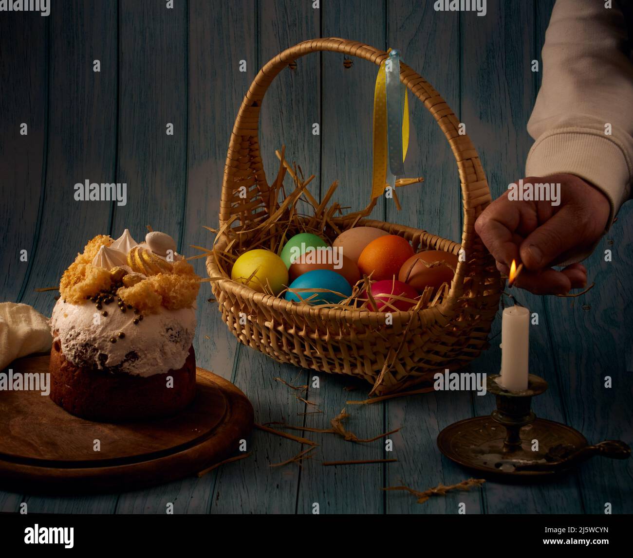 Composition de Pâques. Gâteau de Pâques, un panier avec des œufs de couleur, un porte-bougie en bronze, une main éclairant une bougie avec un fond flou Banque D'Images