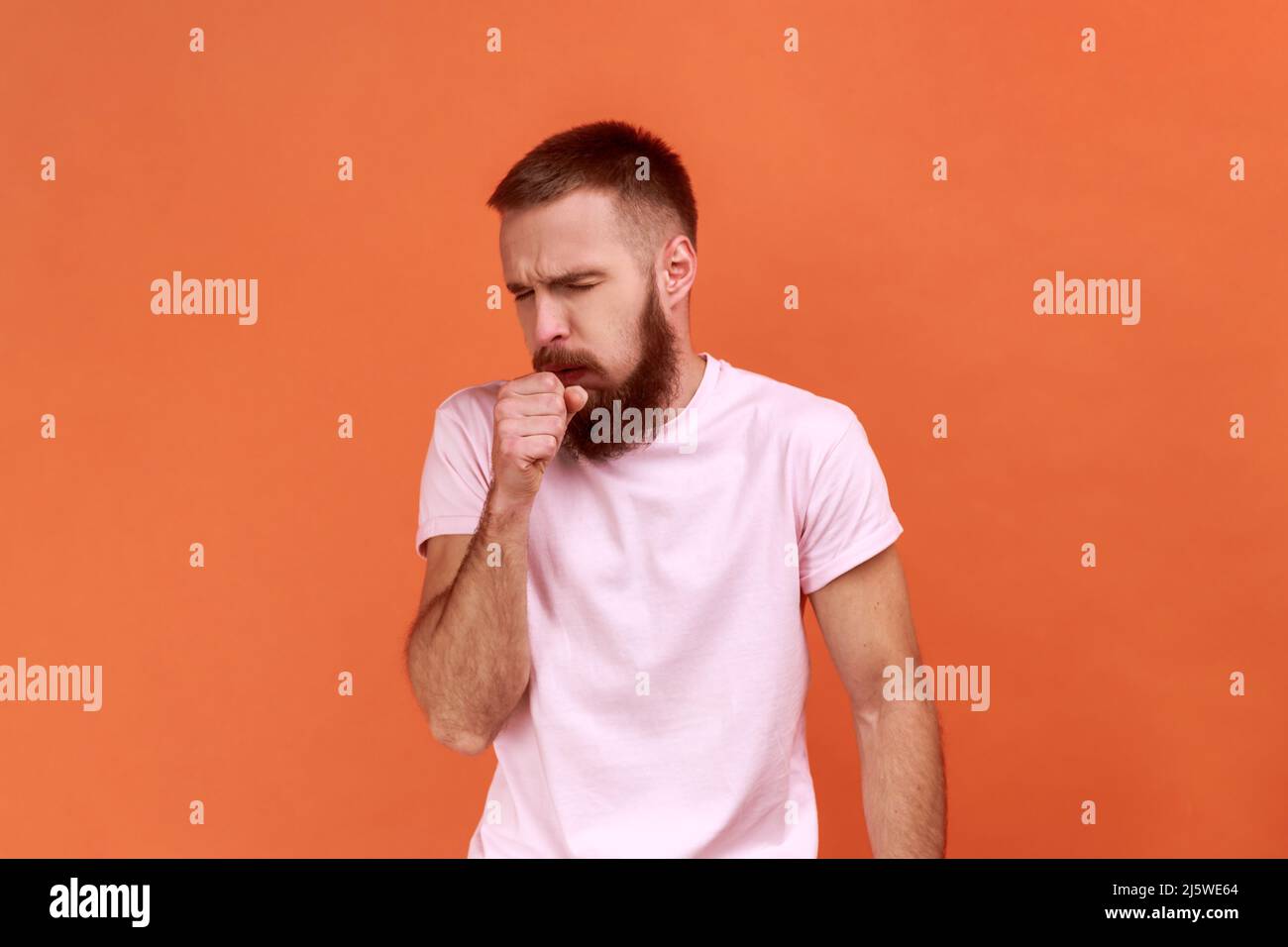 Portrait d'un homme barbu malsain toussant, attrape froid, ayant une température élevée, ayant un symptôme de grippe, portant un T-shirt rose. Studio d'intérieur isolé sur fond orange. Banque D'Images