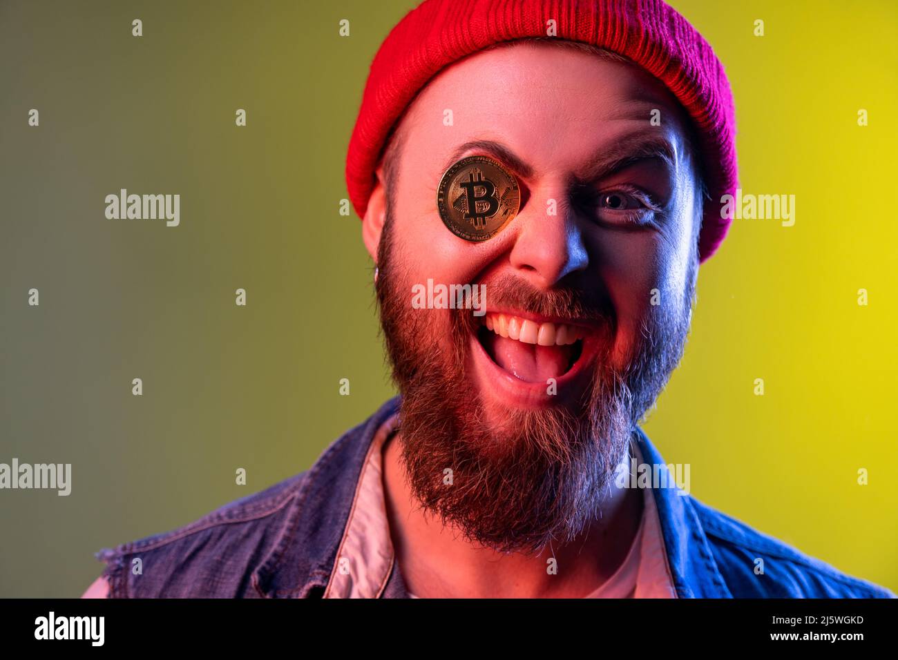 Portrait d'un hipster enjoué et excitant couvrant un œil avec du bitcoin doré, de la crypto-monnaie, portant un bonnet beanie et un gilet en denim.prise en studio en intérieur isolée sur fond coloré de lumière néon. Banque D'Images