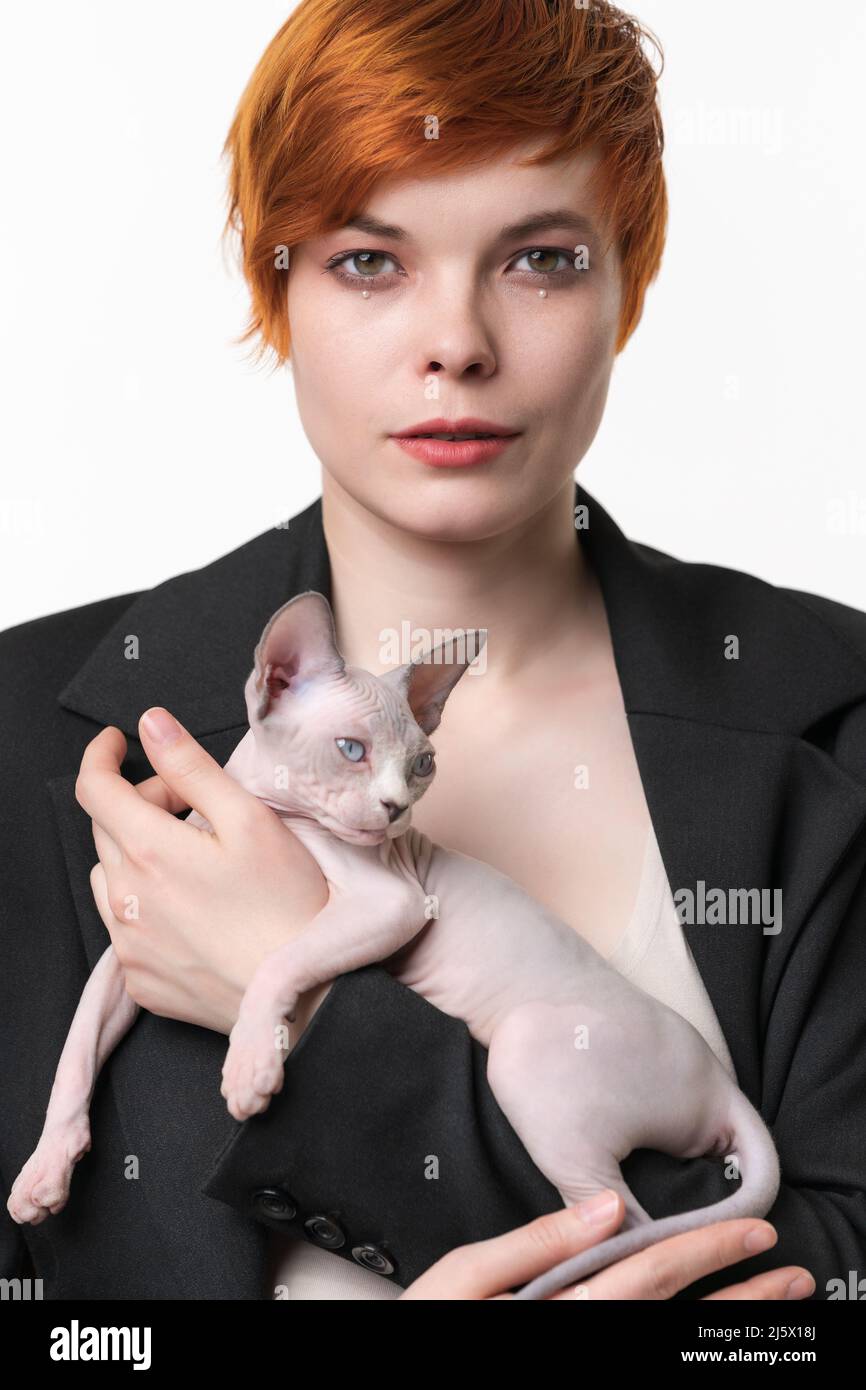 Jeune femme sérieuse en blouson noir tenant entre les mains Canadian Sphynx Cat Jolie femme de 25 ans à tête rouge avec des cheveux courts regardant l'appareil photo Banque D'Images