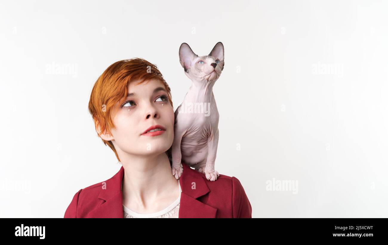 Belle hipster redhead jeune femme avec les cheveux courts regardant vers le haut, vêtu de veste rouge et joueur de chaton de Sphynx assis sur son épaule. Prise de vue en studio Banque D'Images