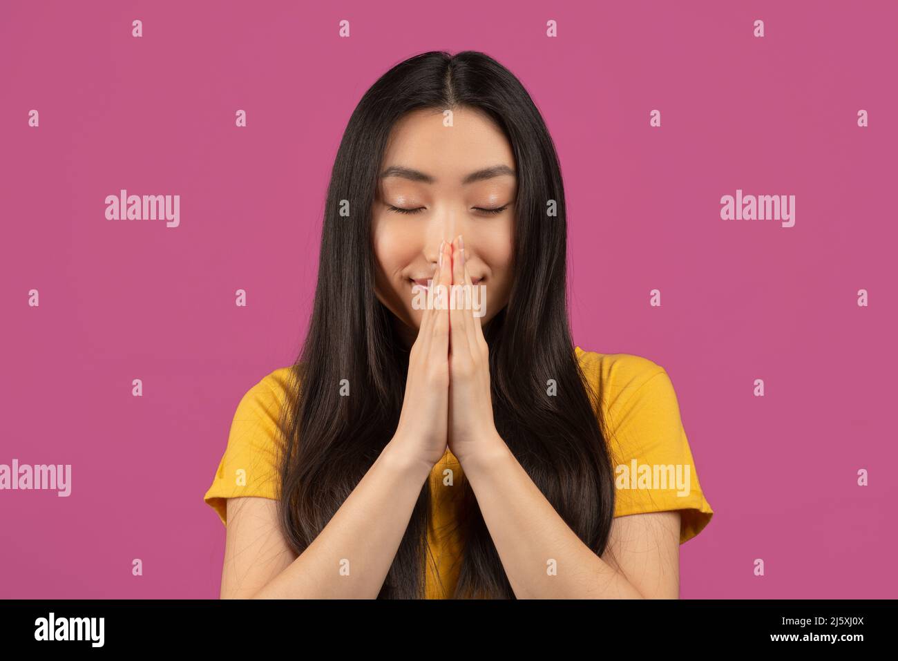 Paisible asiatique dame priant avec les yeux fermés, tenant les mains jointes dans le geste pleurant, debout sur fond violet Banque D'Images