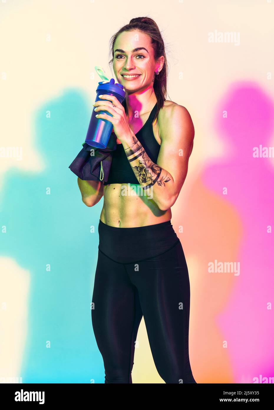 Temps de repos. Une fille sportive avec une serviette sur l'épaule boit de l'eau. Photo de femme musclée en noir sportswear sur fond blanc avec effet RGB colo Banque D'Images