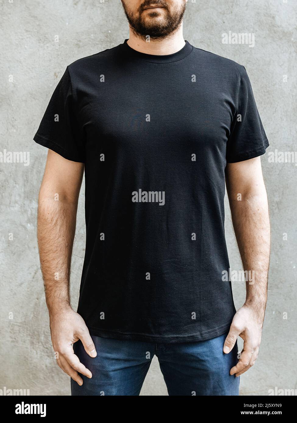 Jeune homme avec une barbe dans un t-shirt noir vide décontracté. Vue avant sur un fond de mur en béton gris clair. Conception et mise en page du t-shirt pour homme pour l'impression. Banque D'Images