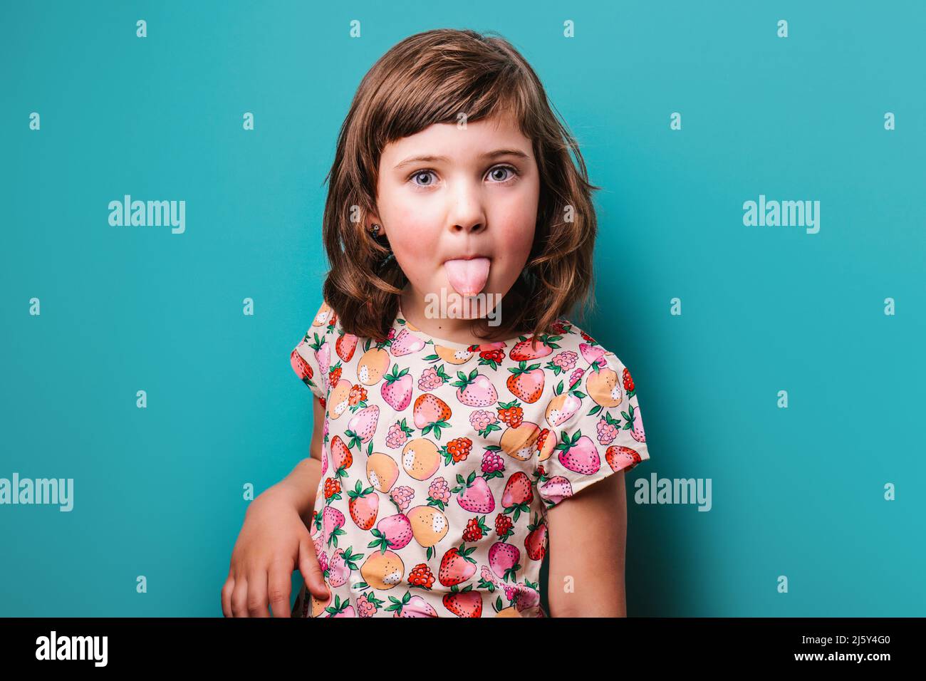 Drôle fille dans l'usure colorée montrant la langue et regardant l'appareil-photo tout en faisant grimace sur fond turquoise en studio léger Banque D'Images