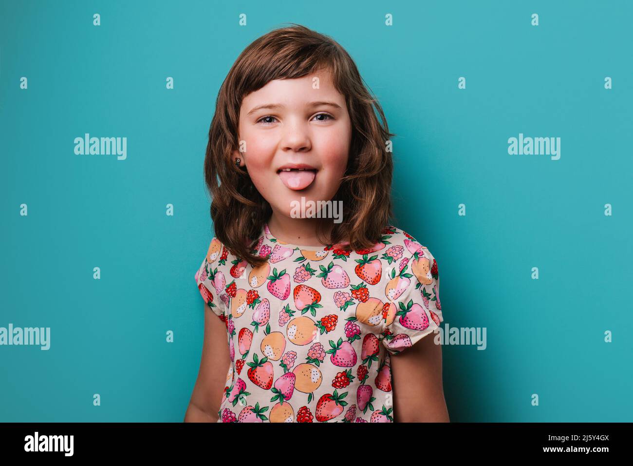 Drôle fille dans l'usure colorée montrant la langue et regardant l'appareil-photo tout en faisant grimace sur fond turquoise en studio léger Banque D'Images