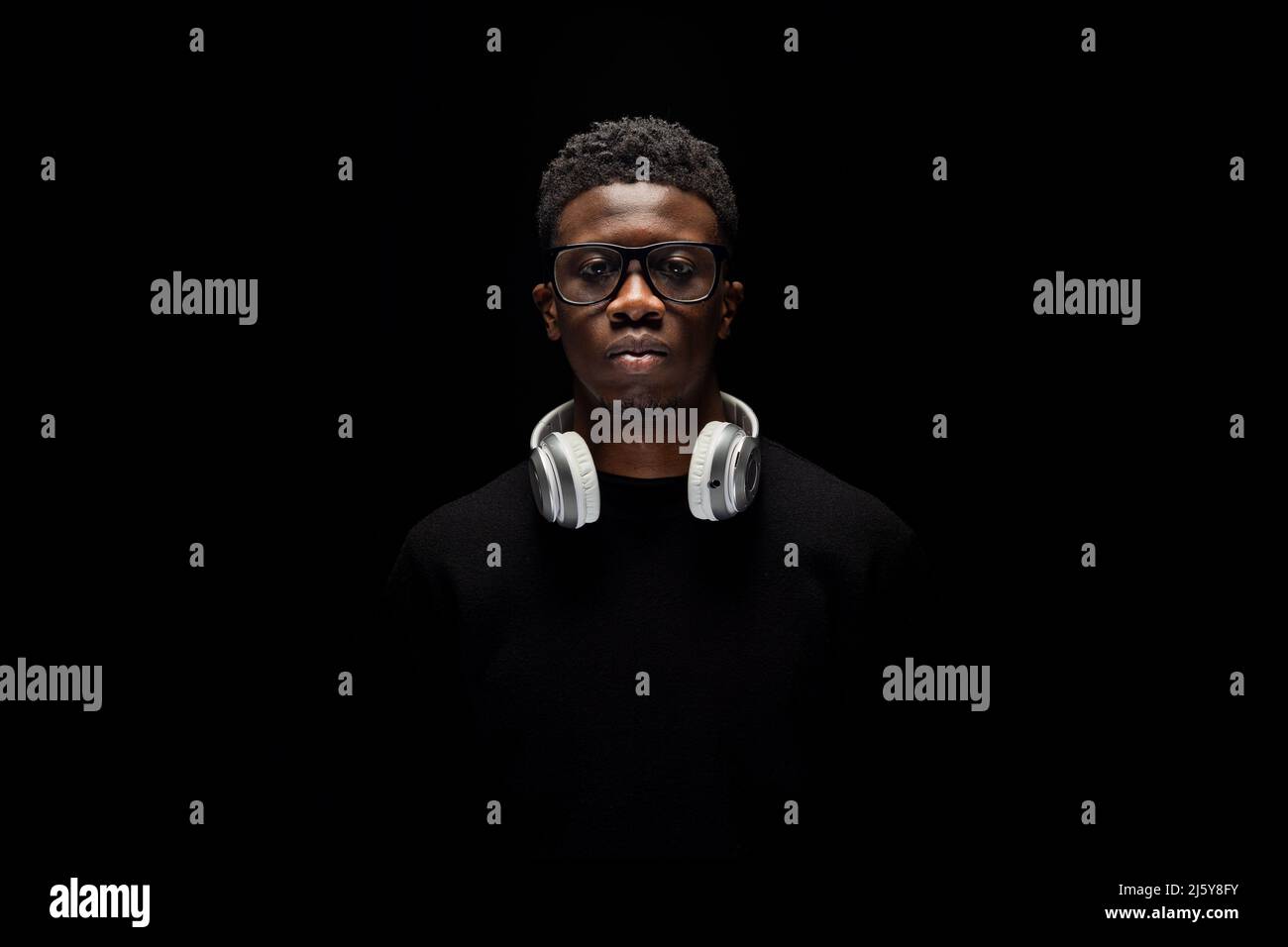 Jeune Afro-américain en lunettes avec casque sur le cou et regardant l'appareil photo sur fond noir Banque D'Images