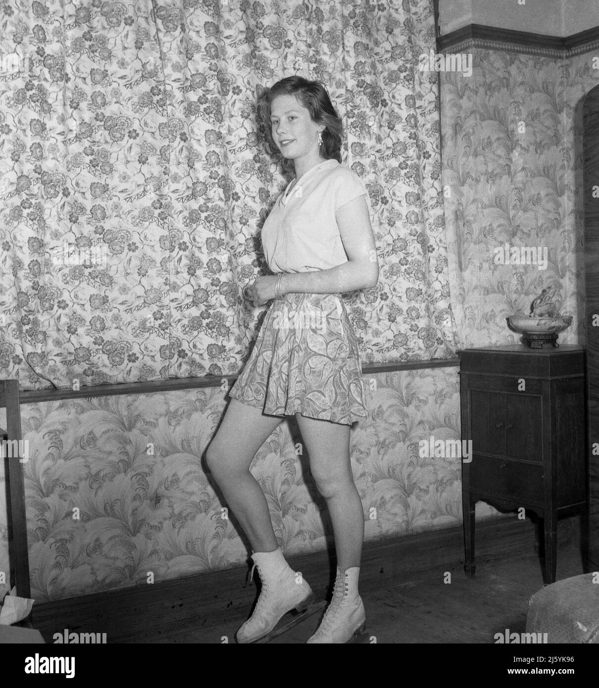 1961, historique, une jeune femme patineuse debout dans une salle de devant pour sa photo dans son sommet de patinage et jupe et patins à glace, Stockport, Angleterre, Royaume-Uni. Sa jupe à motif fleuri est assortie aux rideaux ! Banque D'Images