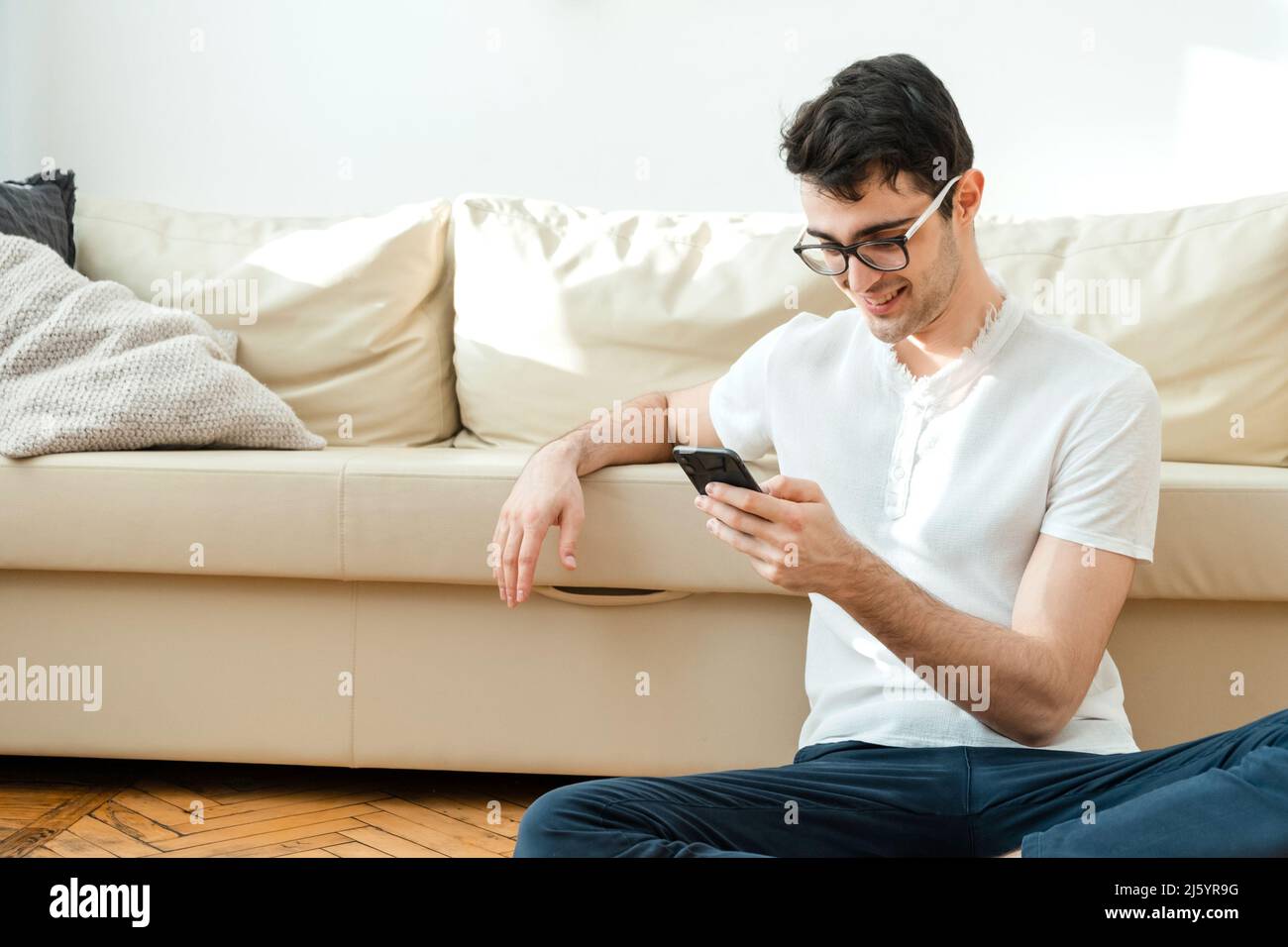 Portrait d'un homme attrayant et souriant dans des vêtements décontractés assis sur le sol près du canapé dans le salon et regardant les nouvelles à l'aide d'un ph mobile Banque D'Images