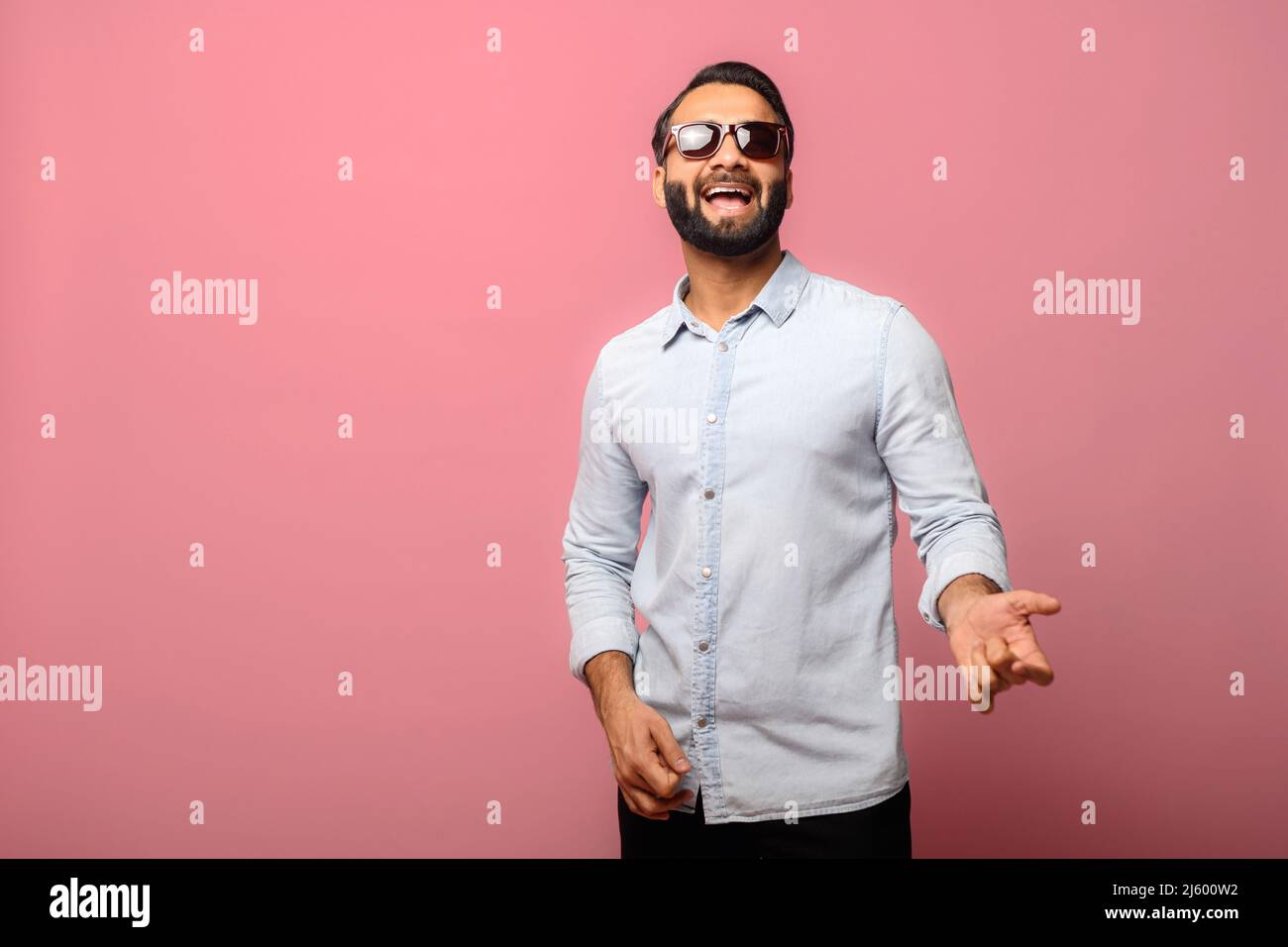 Joyeux Indien en lunettes de soleil prtend guitariste joueur, excité hispanique gars dans décontracté Jean chemise aime jouer sur la guitare imaginaire, a un plaisir, isolé sur rose Banque D'Images