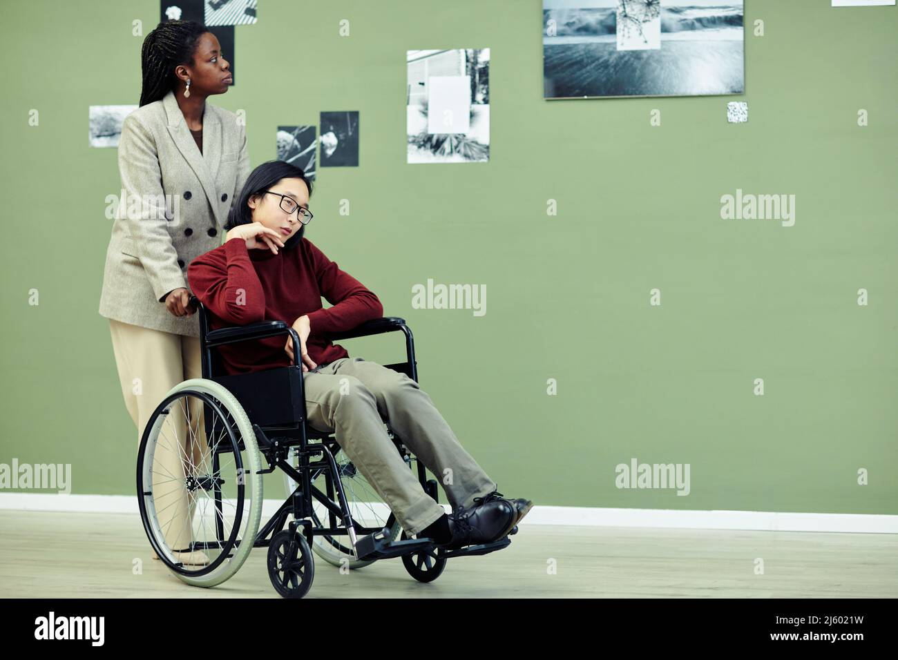 Une jeune femme afro-américaine et son amie asiatique masculine handicapée en fauteuil roulant visitent ensemble l'exposition de photographie moderne Banque D'Images