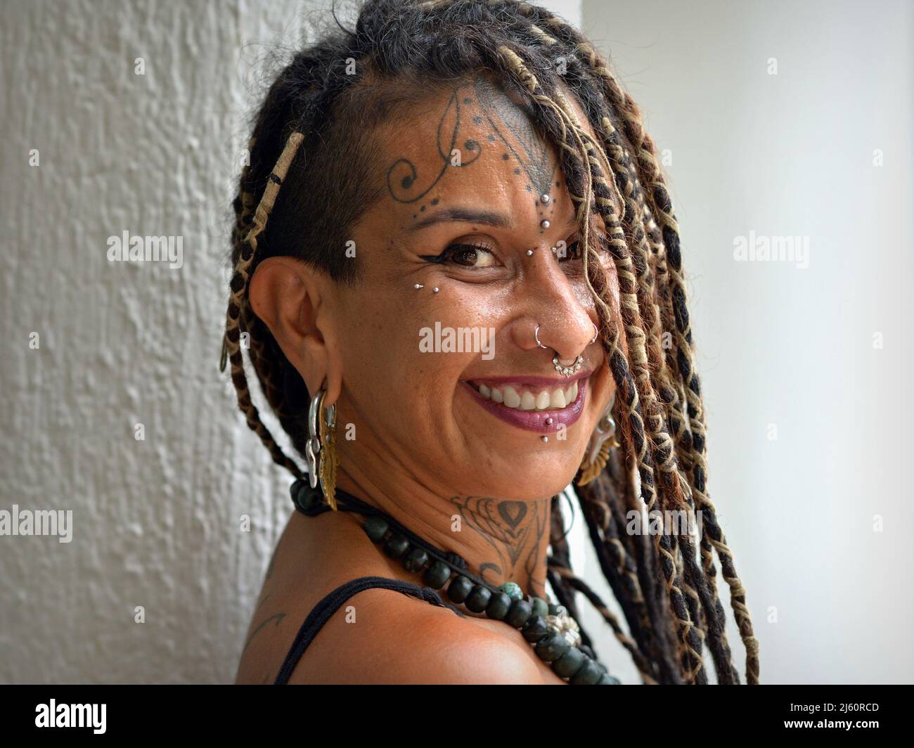 Une jeune femme mexicaine alternative attrayante avec de longues tresses rasta, un piercing facial d'art corporel et des sourires de tatouage positivement optimiste au spectateur. Banque D'Images