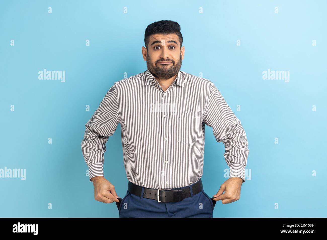 Un homme d'affaires malheureux déprimé avec une barbe montrant des poches vides de son pantalon en denim, regardant l'appareil photo avec tristesse, faillite, portant une chemise rayée. Studio d'intérieur isolé sur fond bleu. Banque D'Images