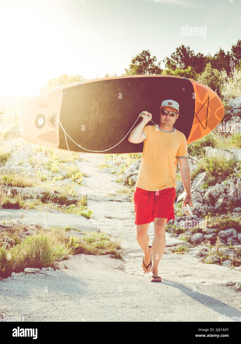 surfeur mâle transportant la planche sur l'épaule et marchant sur un petit chemin; situation de contre-jour avec beau coucher de soleil en soirée Banque D'Images