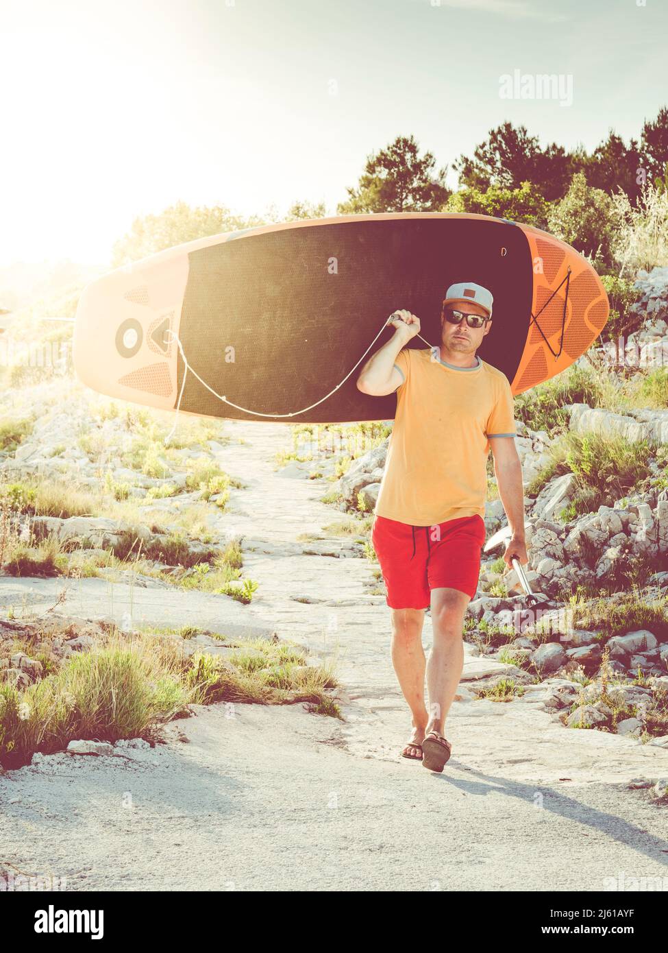 surfeur mâle portant un paddle-dessus de l'épaule et marchant sur un petit chemin; situation de contre-jour avec beau coucher de soleil en soirée Banque D'Images