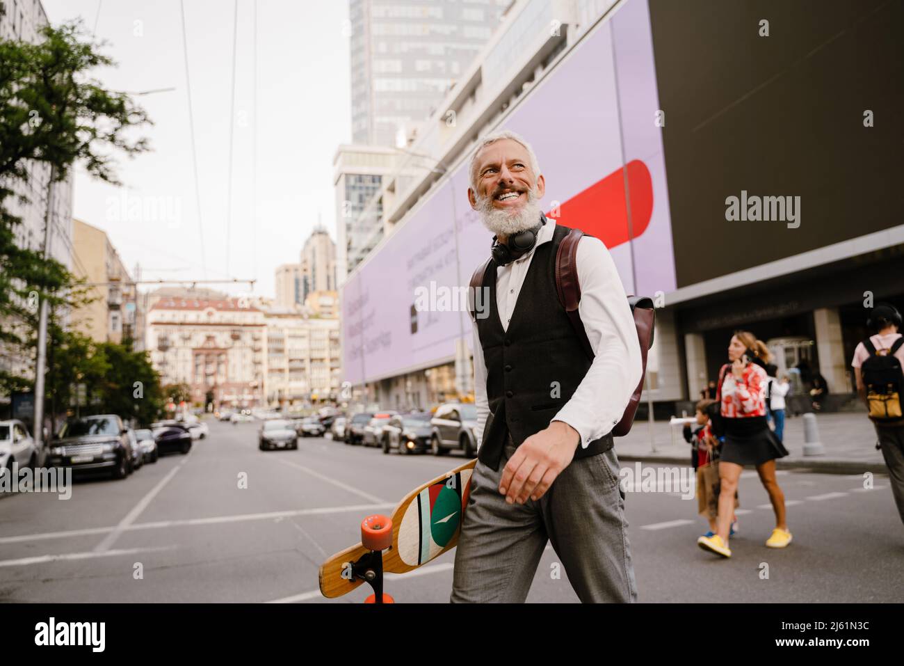 Homme gris mûr souriant tout en tenant avec un skateboard tout en marchant dans la rue de la ville Banque D'Images