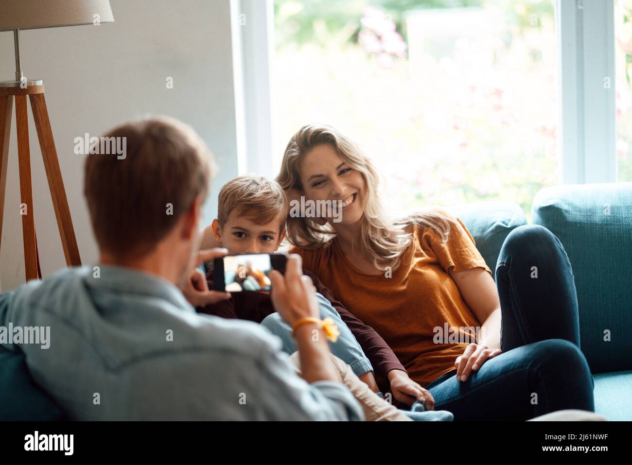 Bonne femme blonde et fils photographiés par l'homme à travers le smartphone dans le salon Banque D'Images