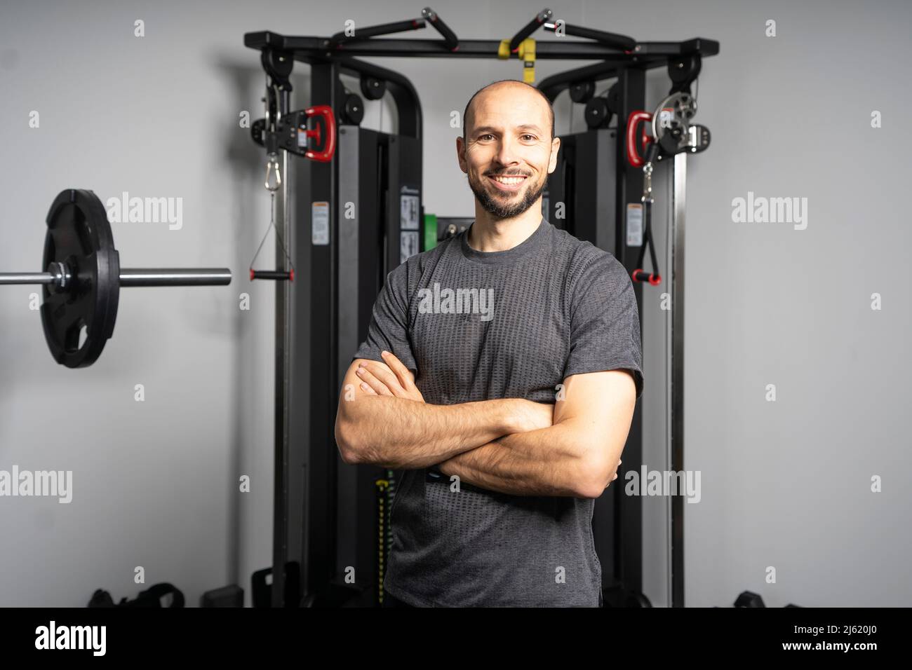 Instructeur de fitness souriant debout avec les bras croisés devant l'équipement d'exercice à la salle de gym Banque D'Images