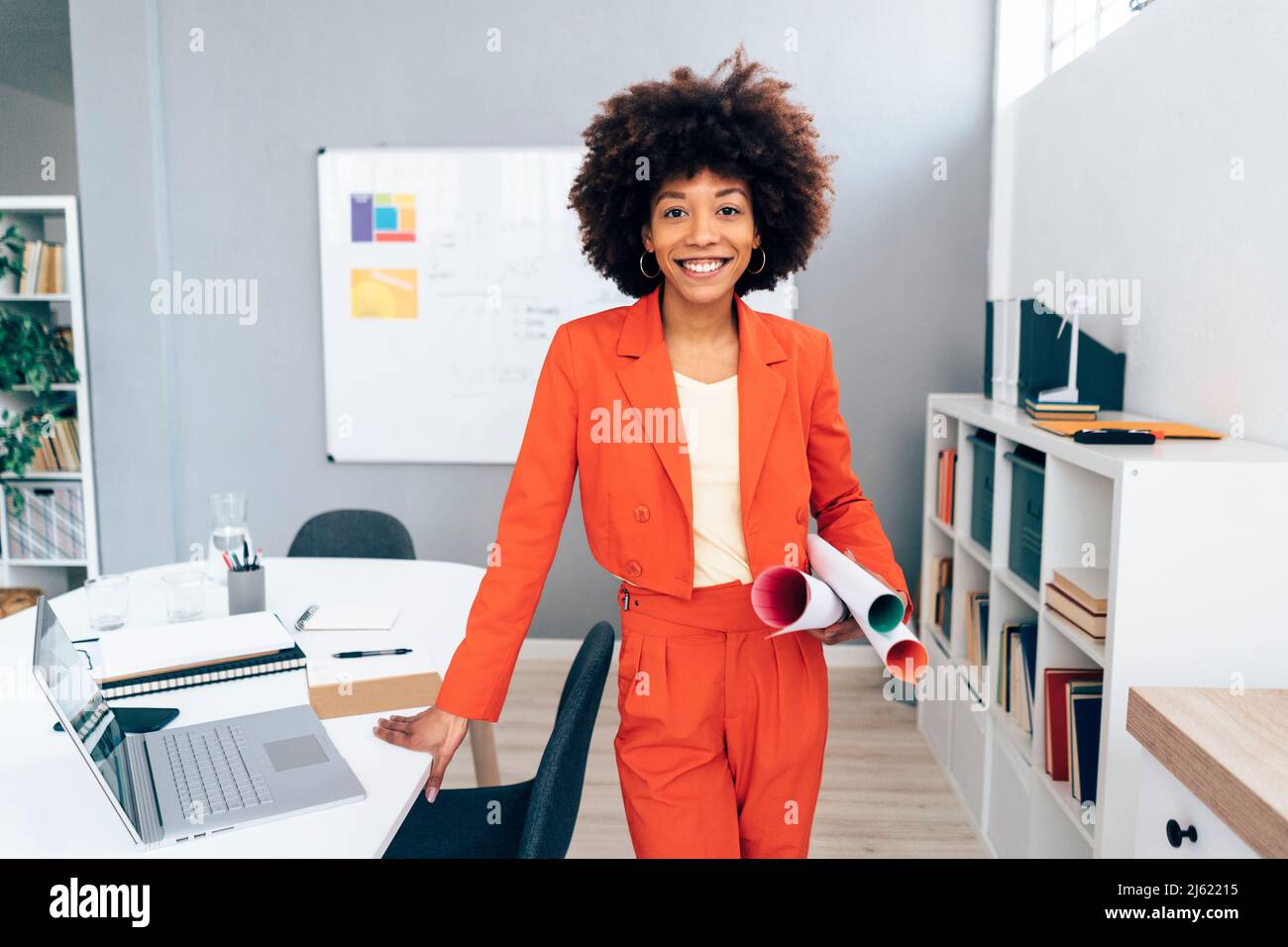 Femme d'affaires souriante avec une coiffure afro tenant un plan au bureau Banque D'Images