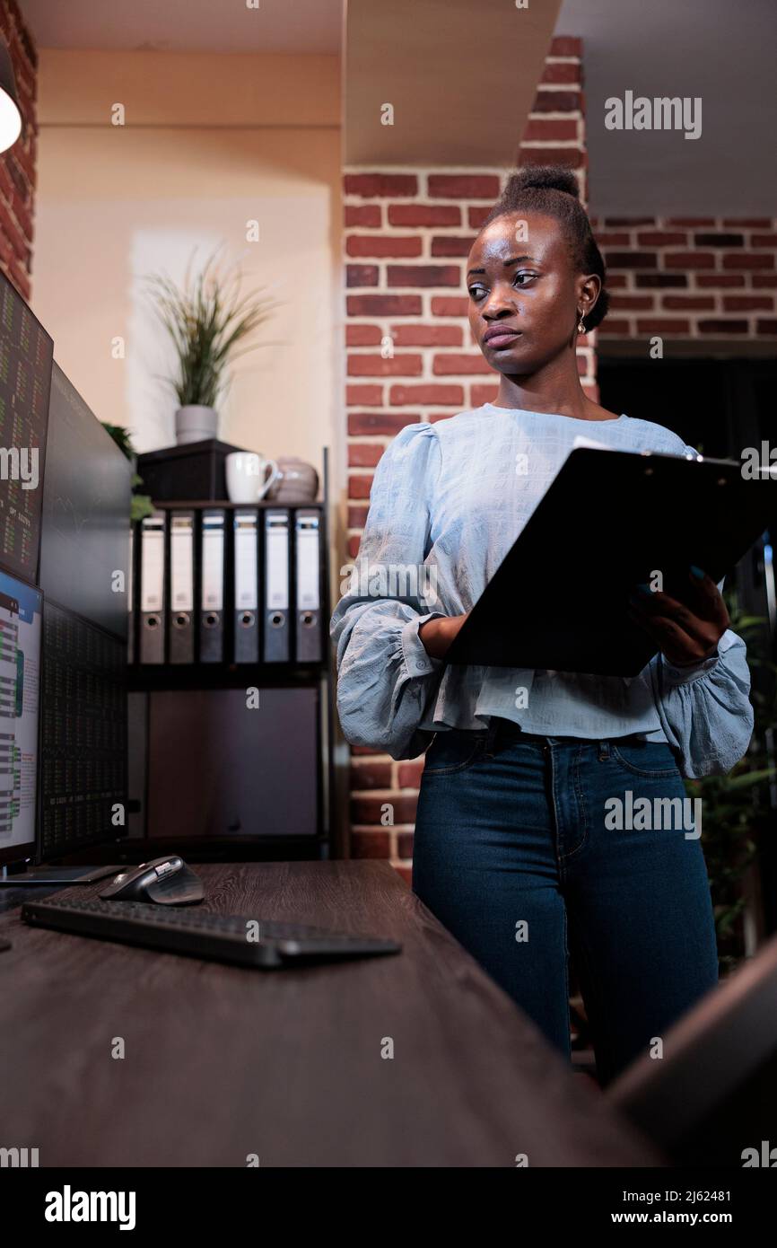 Société d'investissement afro-américaine analyste financier professionnel avec presse-papiers analysant les données du marché en temps réel. Agent d'agence de courtage debout devant un poste de travail à plusieurs moniteurs avec presse-papiers. Banque D'Images