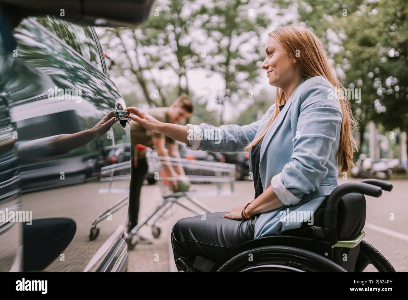 Jeune femme souriante assise en fauteuil roulant et ouvrant la porte de voiture Banque D'Images