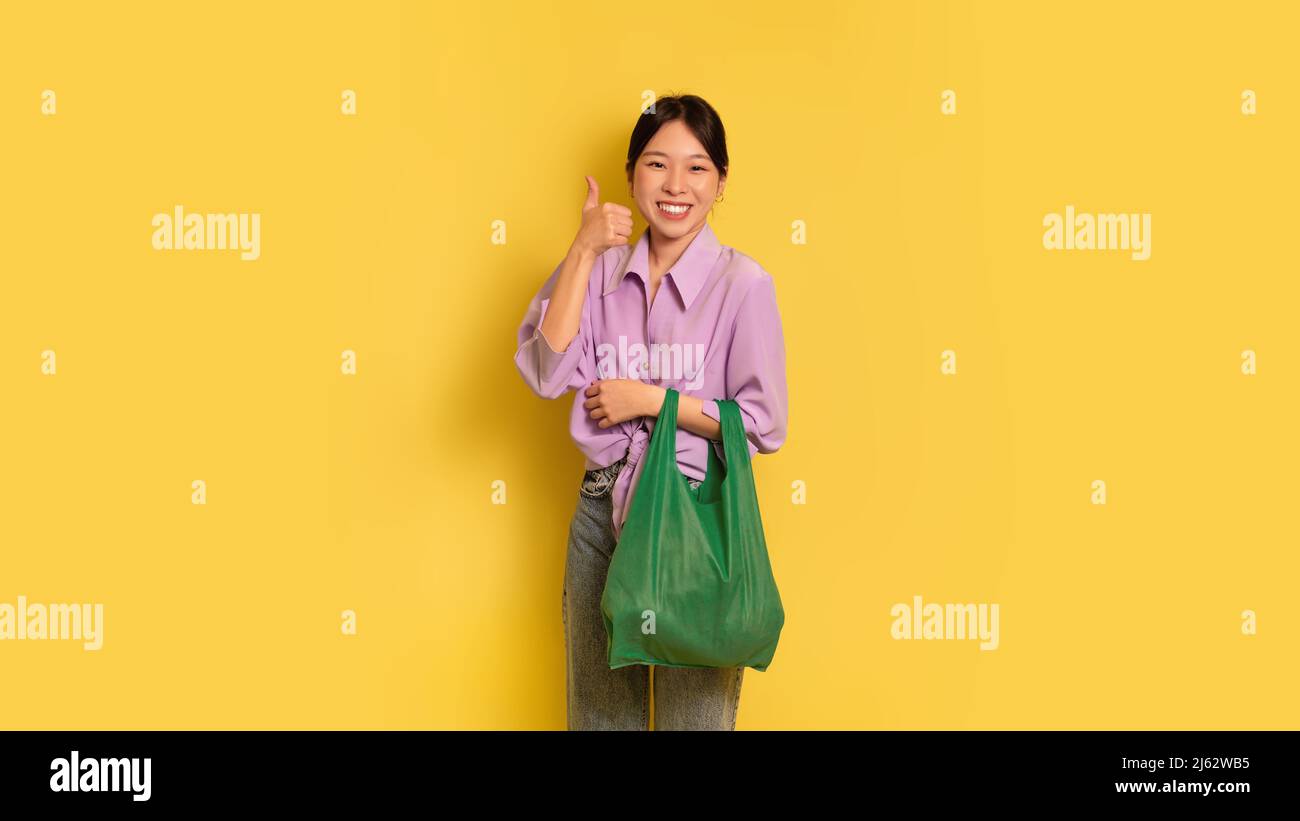 Une femme asiatique positive tenant un sac écologique réutilisable et montrant un geste de pouce vers le haut, recommandant un shopping sans gaspillage Banque D'Images