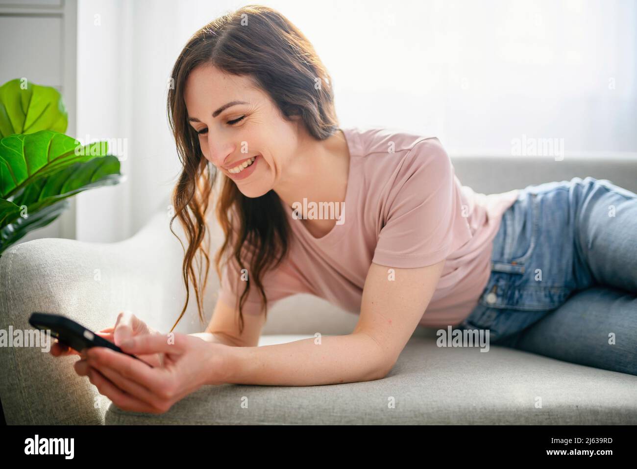 sympathique fille à cheveux longs utilisant un téléphone portable assis à la maison sur un canapé Banque D'Images