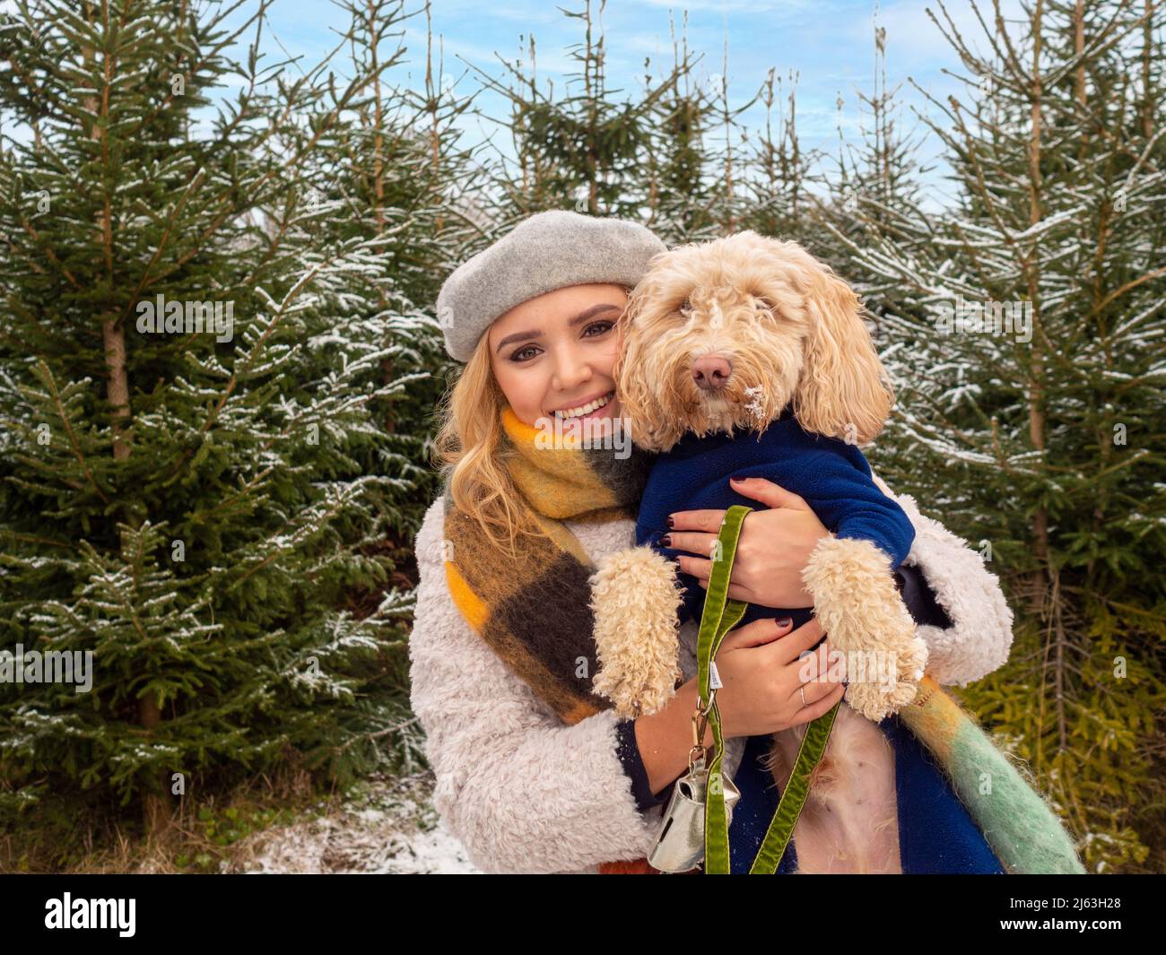 Jeune femme caucasienne vêtue d'un chapeau et d'un manteau chauds, tenant un chien posant dans des arbres de Noël enneigés. Banque D'Images