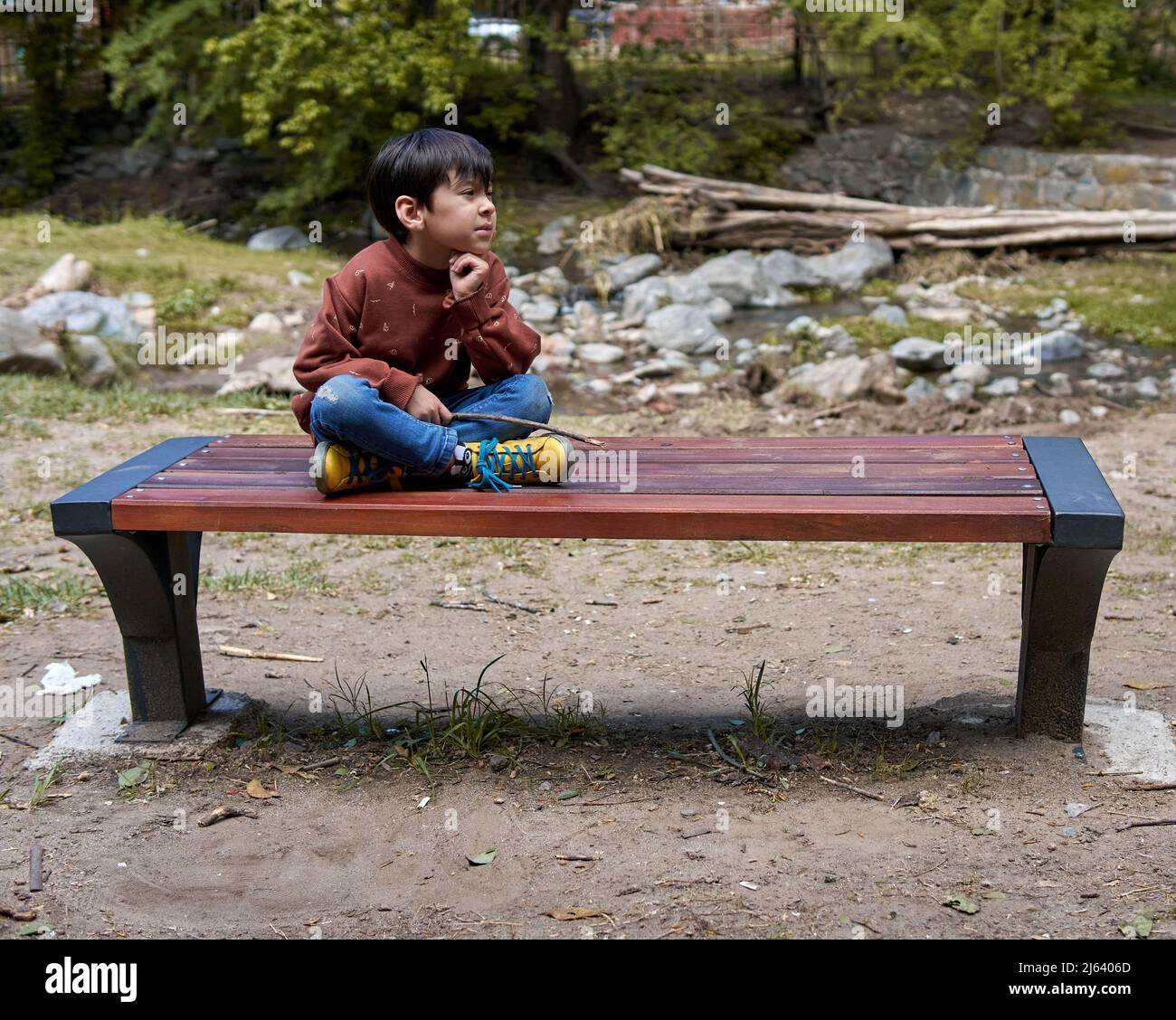 Petit garçon Latino solitaire pensif assis sur un banc de fer et de bois à côté d'un ruisseau dans des vêtements urbains regardant le paysage. Horizontal et espace de copie. Banque D'Images