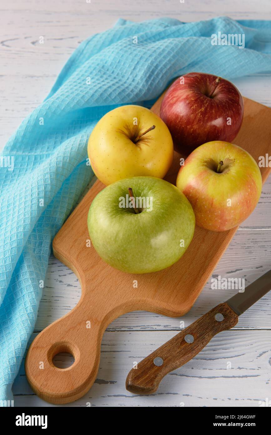Les pommes multicolores reposent sur une planche en bois sur un fond en bois blanc. À proximité se trouvent une serviette bleue et un couteau. Banque D'Images