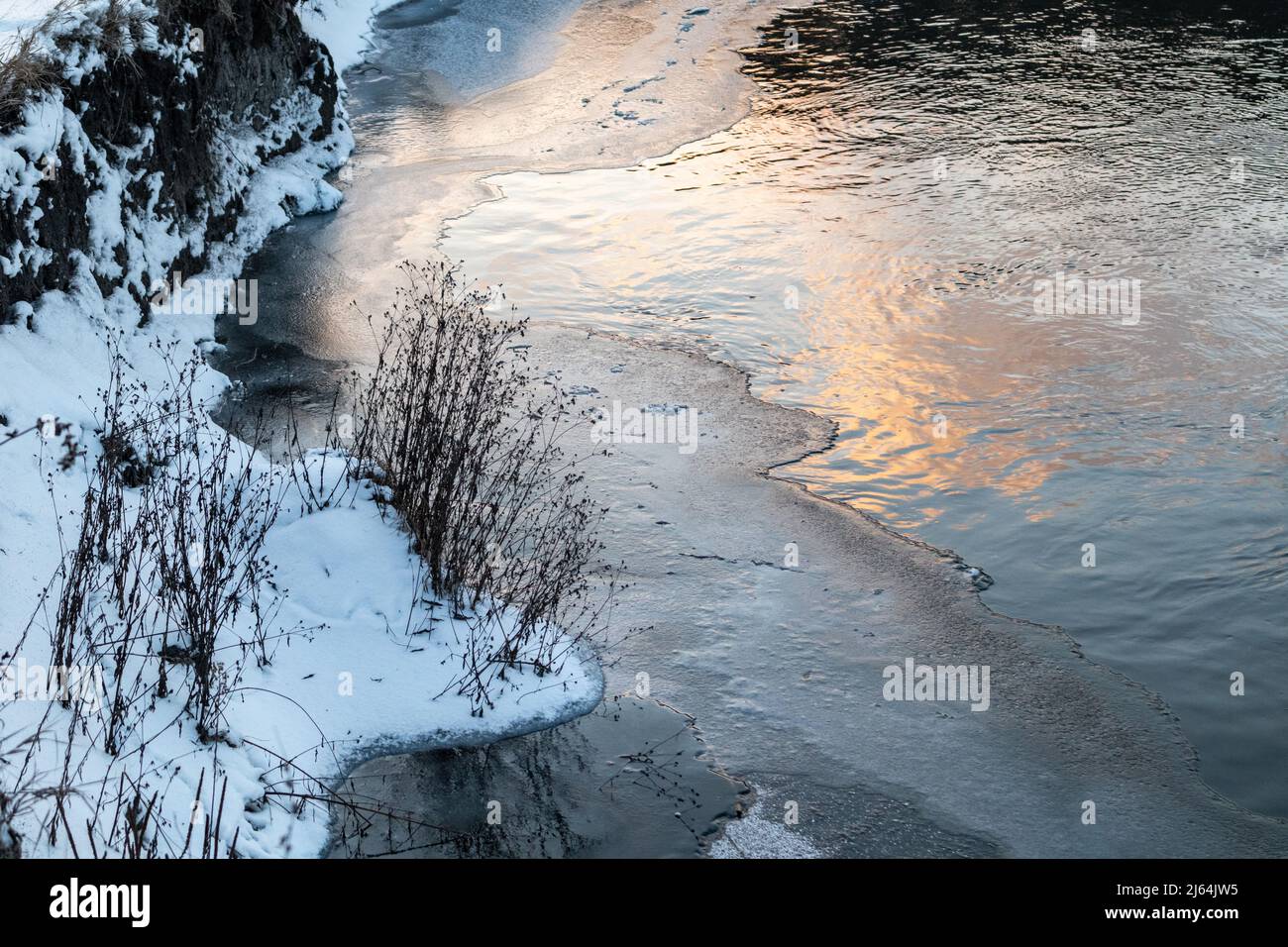 Débit d'eau sur le lac recouvert de neige en gros plan. Vue d'hiver sur le rivage de la rivière gelée froide avec de l'herbe sèche et des nuages de coucher de soleil réfléchissent Banque D'Images