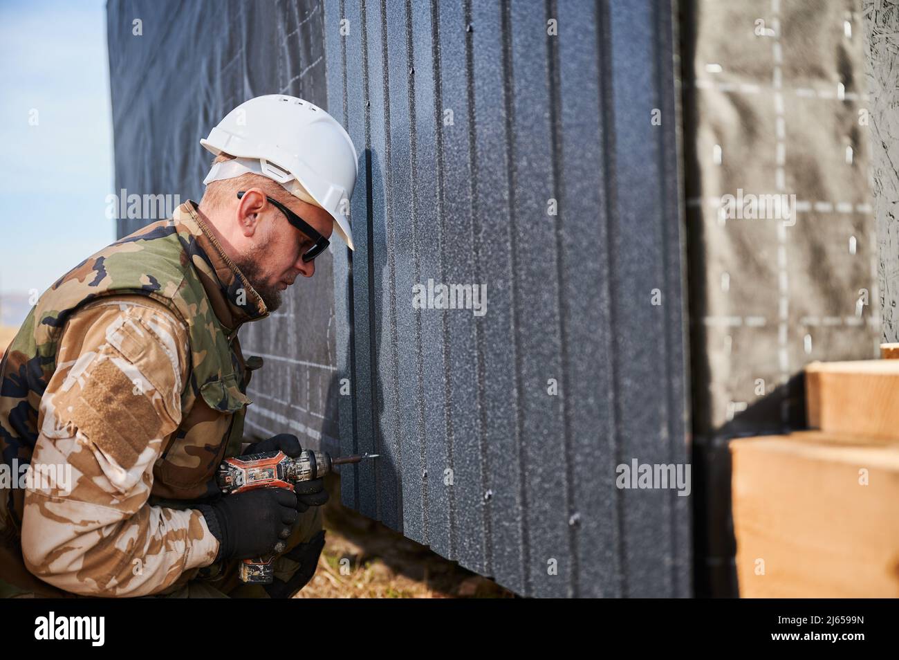 Un constructeur mâle installe une tôle de fer ondulée noire utilisée comme façade de future maison. Homme ouvrier bâtiment maison en bois cadre. Conception de la menuiserie et de la construction. Banque D'Images