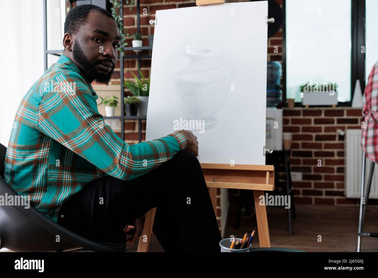 Portrait d'un élève souriant assis devant un vase en toile blanche à l'aide d'un crayon graphique pendant la leçon d'art en studio de créativité. Jeune artiste développant des compétences artistiques pour la croissance personnelle Banque D'Images