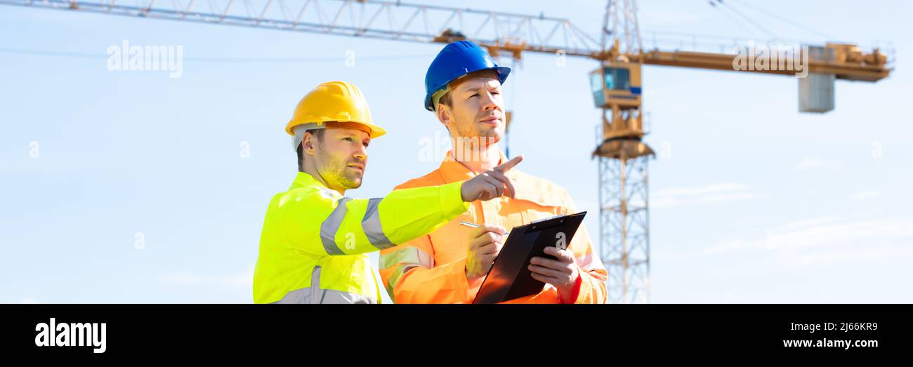 Deux jeunes ingénieurs masculins debout en face de la grue mécanique travaillant sur le chantier de construction Banque D'Images