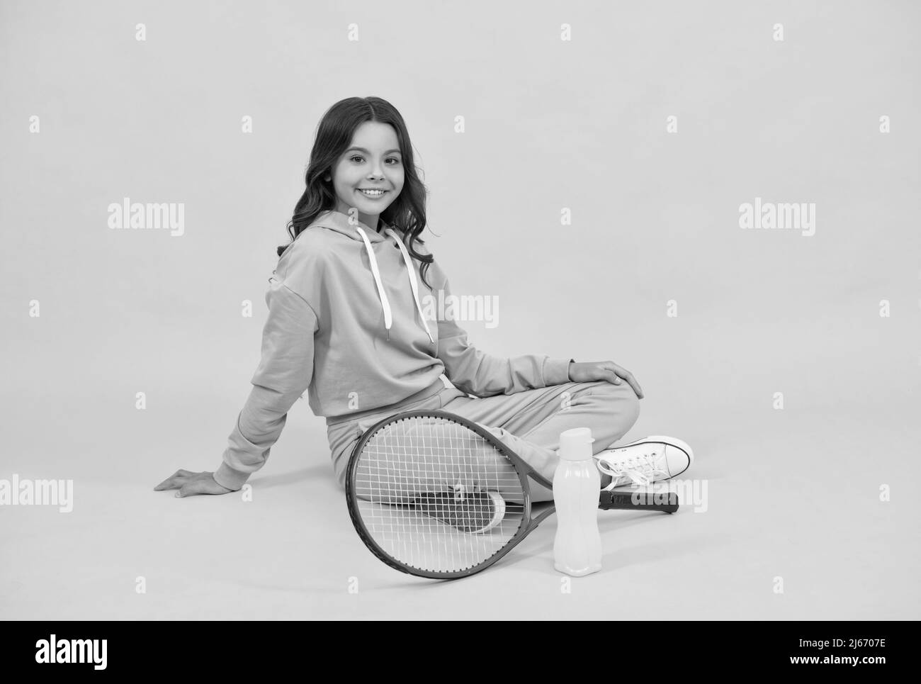 un enfant joyeux s'assoit dans des vêtements de sport avec une raquette de squash et une bouteille d'eau sur fond jaune, santé Banque D'Images