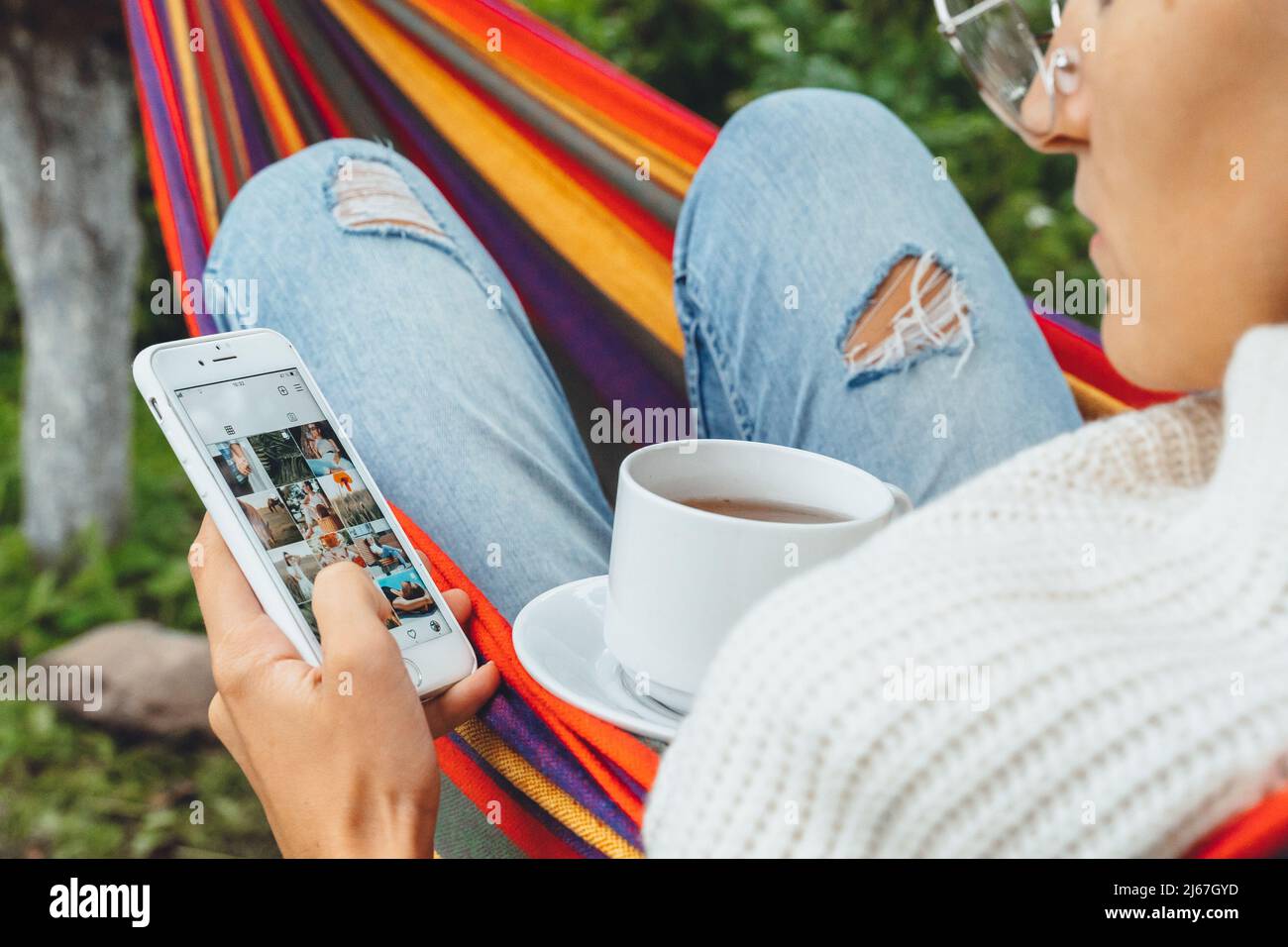 Joli créateur de contenu dans hamac utiliser smartphone regarder l'écran de chat dans les médias sociaux boire thé lire les nouvelles en ligne navigation photo en vacances, vue arrière Banque D'Images